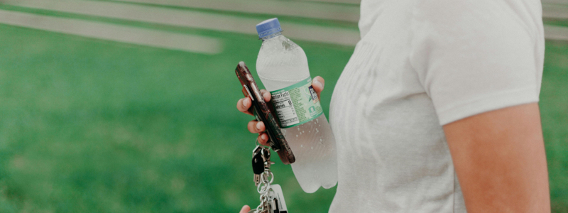 Close crop into a woman's torso. She is holding a plastic water bottle, cell phone, and keys, as she walks across a grassy field.