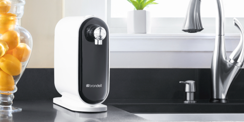 Brondell Cypress Three-Stage Countertop Water Filtration System sits atop a black quartz countertop next to the sink with a decorative vase filled with lemons.